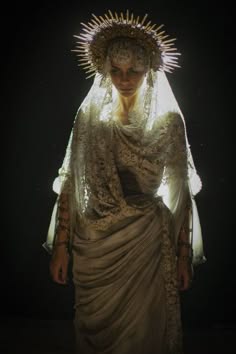 a woman in a white lace dress with a gold crown and veil, standing in a dark studio, looking down with a mysterious and alluring expression. she is surrounded by soft light that highlights her features and clothing.
