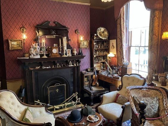 this is an image of a victorianstyle living room. the walls are covered in a deep red patterned wallpaper and the furniture is made of dark wood. a fireplace is on the left side of the image with a mirror above it. there are two armchairs in the foreground, and a small table in the center of the room. a window is on the right side of the image, and a lamp is lit on the table next to the window. the room is decorated with various items, including candlesticks, a clock, and a collection of small figurines.  the lighting is a mix of natural light from the window and artificial light from the lamp.
