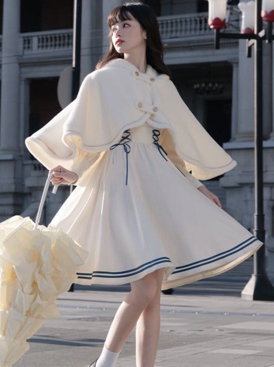 a young woman in a white dress and matching cape stands on a city street. she is holding a yellow umbrella and looking to the side. the photo is taken from an eyelevel perspective and features natural daylight.