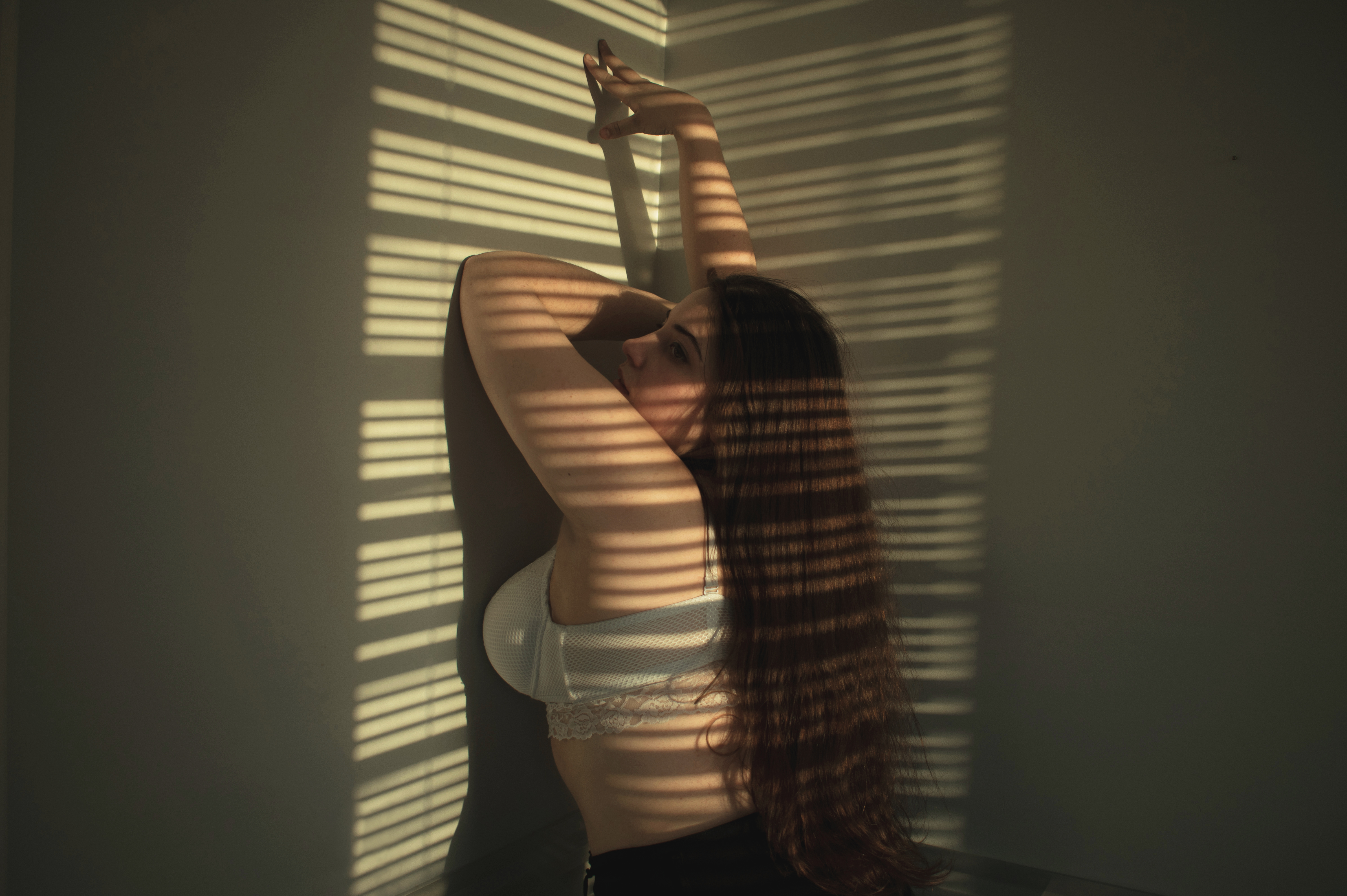 a woman in a bra is leaning against a corner wall, with light from window blinds creating shadows on her body and the wall. she has her arms raised.