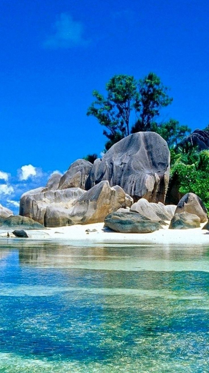 a stunning tropical beach scene with clear, turquoise water lapping at the shore. large granite rocks dot the white sand, creating a dramatic backdrop for the idyllic scenery. the sky is a vibrant blue with wispy white clouds, and lush green foliage adds to the lush atmosphere. the scene is bathed in sunlight, making the colors of the water and sand even more vibrant. it is a perfect example of the beauty and serenity of the natural world.