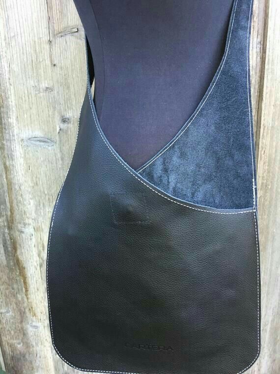 a black leather shoulder bag is displayed on a mannequin. the bag features white stitching and a textured exterior. the inside appears to be suede. the bag hangs against a wooden background.