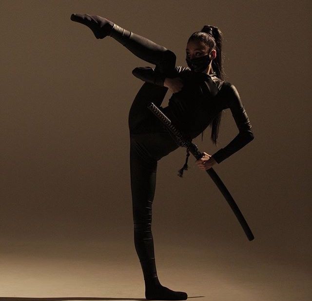 a woman is holding a katana sword in one hand and bending backwards,  wearing all black outfit. she is wearing a black mask over her nose and mouth. the background is brown.  she is likely practicing martial arts.