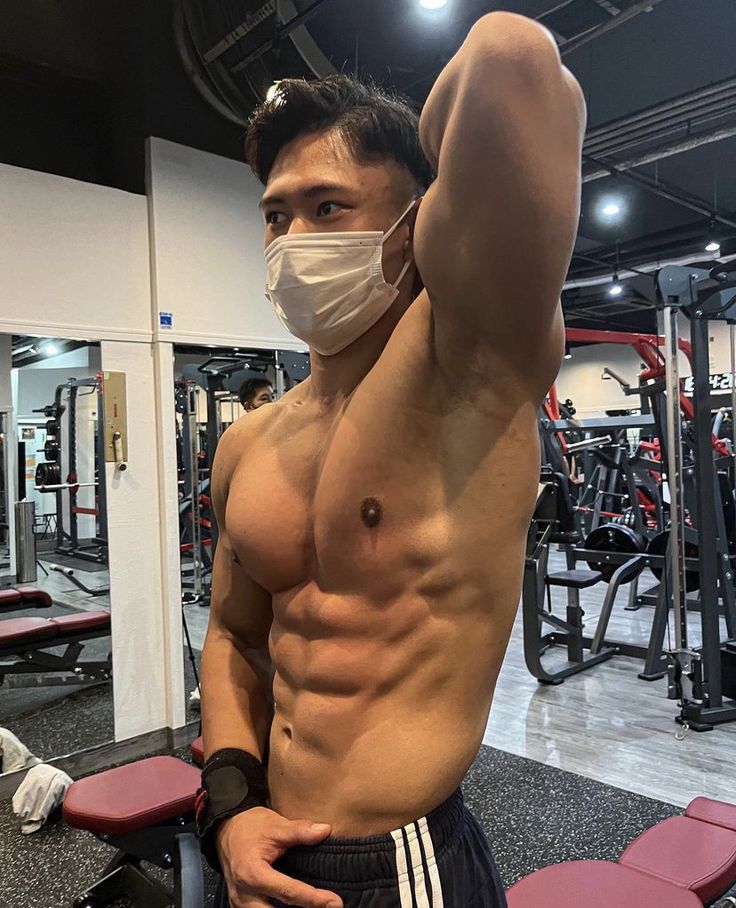 a muscular asian man wearing a face mask is posing in a gym, showing off his sixpack abs. he has his arm raised and is looking towards the left side of the frame.  the background shows gym equipment. the image focuses on his physique.