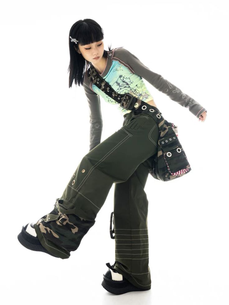 a fullbody shot of a young woman, possibly a fashion model, posing in a stylish outfit against a clean white background. she is wearing a longsleeved, graphic tee, olive green cargo pants with camouflage accents, and chunky black boots.  a crossbody bag with a camouflage pattern is slung over her shoulder. her black hair is styled simply with a hair clip. the overall style is edgy, alternative, and leans toward japanese streetwear or harajuku fashion. the image is welllit and captures the details of her attire.
