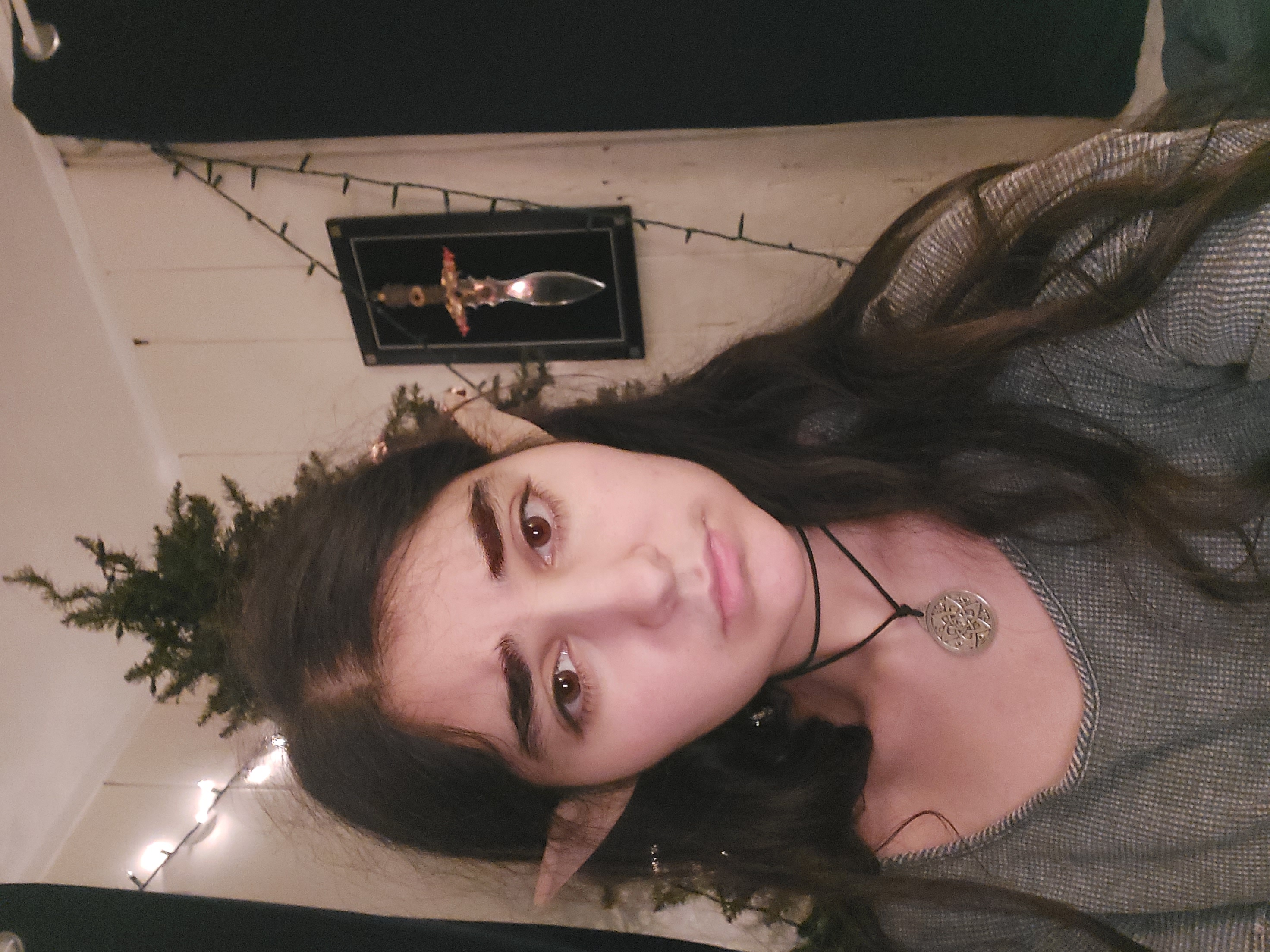 closeup selfie of a young woman with long dark hair wearing elf ears, a grey sweater, and a necklace with a circular pendant. she has a serious expression on her face.  a small christmas tree with lights is visible in the background, along with a framed dagger on the wall. the setting appears to be a bedroom.