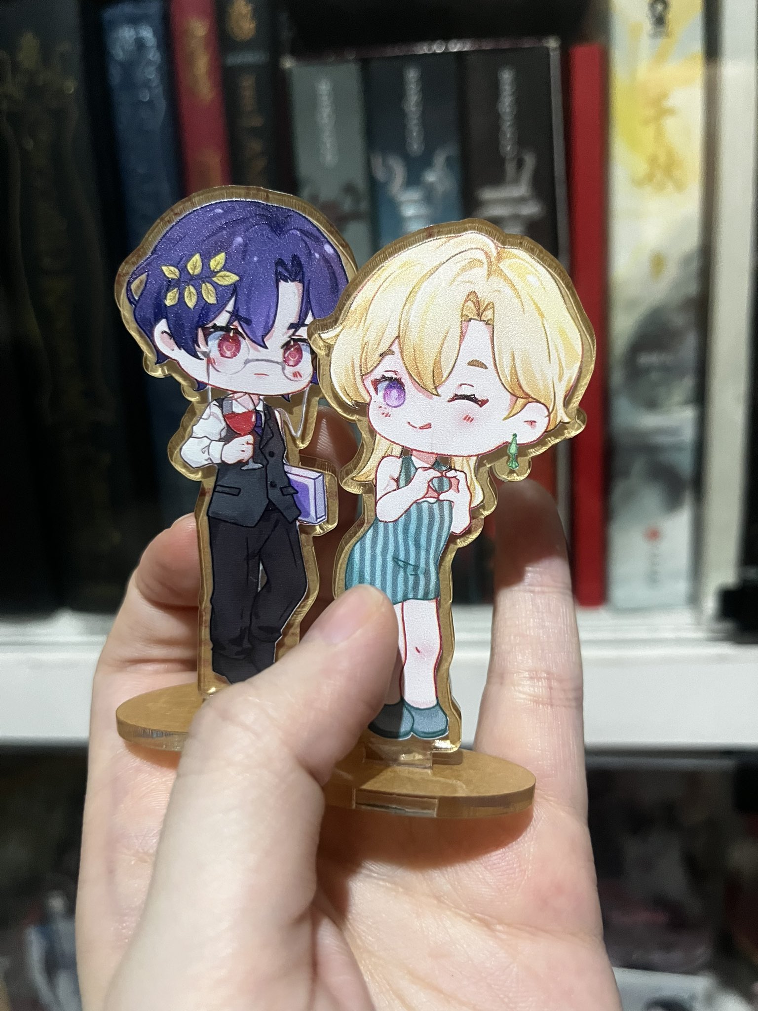 a hand is holding a cute acrylic stand with two anime characters on it.  a boy with purple hair, wearing a black suit and glasses is holding a book and a glass of red liquid. he is standing next to a girl with blonde hair, wearing a green dress and a red bow. the characters are standing in front of a bookshelf with several books. the stand is made of wood and has a golden frame. the hand holding the stand is also on a background with a shelf.  the photo is taken in a closeup shot.