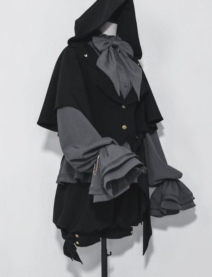 a gothic lolita outfit is displayed on a mannequin. the outfit consists of a black hooded cloak or cape, a grey blouse with a large bow, and a black ruffled skirt or shorts. the blouse features long grey sleeves with multiple layers of ruffles at the cuffs. the outfit is mostly black and grey. a black sash or ribbon is tied around the waist. the photograph has a studiolike setting with a plain white background.