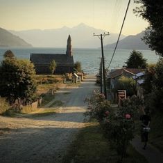 the image shows a small village nestled on the shore of a lake, with towering mountains in the distance. a quaint church with a steeple stands prominently on the left side of the road that leads through the village. the road is unpaved and dirt, suggesting a sense of rural tranquility.  the presence of lush green trees adds to the serene ambiance. a lone person can be seen walking along the roadside, their back turned to the camera. the soft, natural lighting, likely from the setting sun, bathes the scene in a warm, ethereal glow, emphasizing the peaceful and picturesque nature of this remote location.