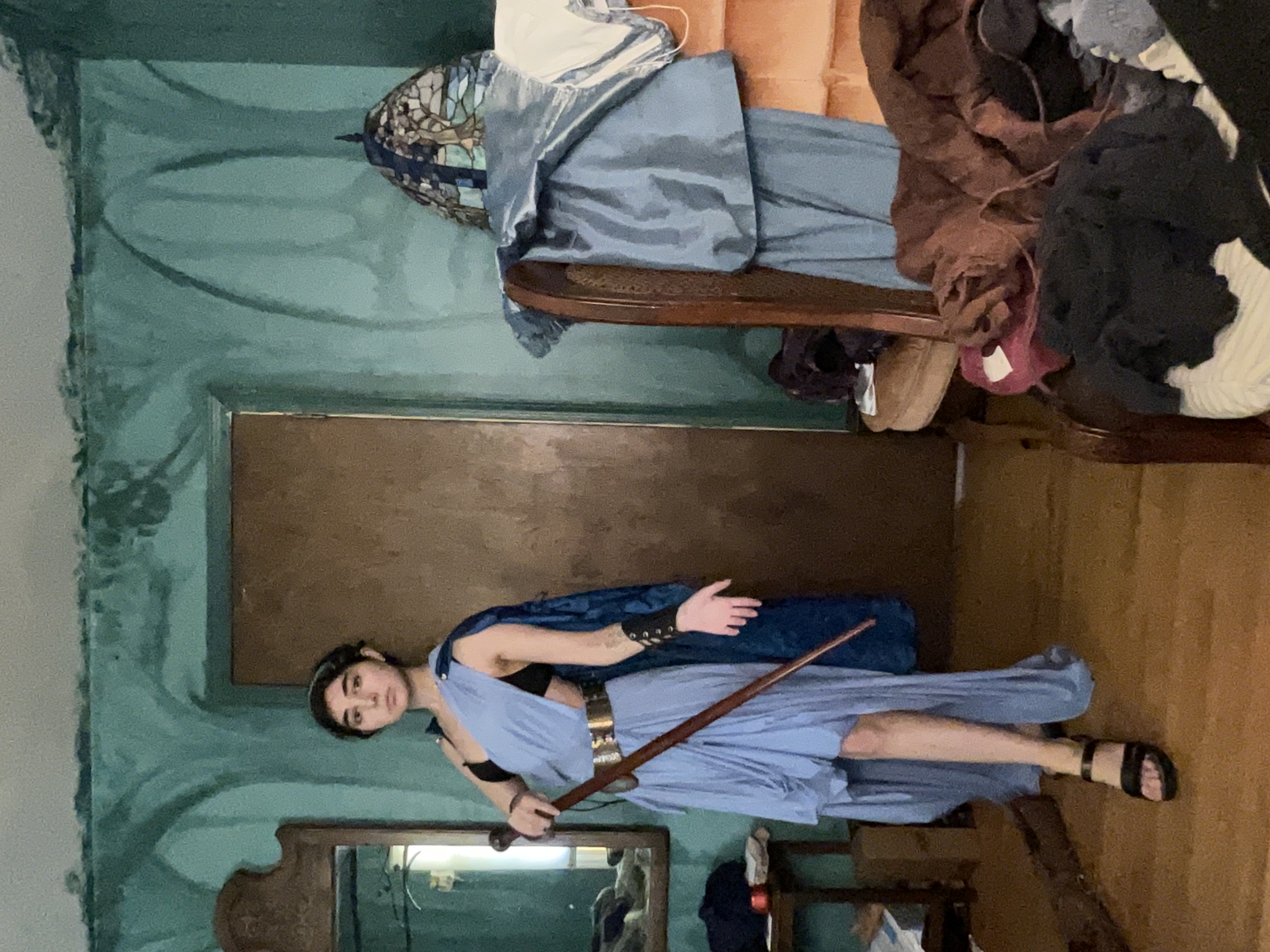 young woman is posing in a cosplay costume. she's wearing a light blue, flowing dress reminiscent of ancient greek attire, paired with a darkcolored head band and sandals. she holds a staff and seems to be portraying a warrior character. the background shows a messy room with teal walls, a vintagestyle mirror, and various furniture items scattered around.  the overall impression is a casual cosplay photo taken in a less than pristine setting.