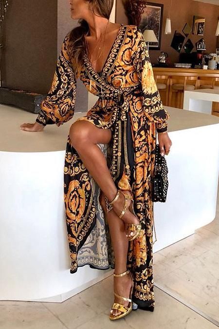 a woman is posing in a stylish maxi dress with a baroque print. the dress is black and gold and has long sleeves and a wrapstyle vneck. she is wearing gold high heels and carrying a small black clutch. the setting appears to be a modern home, and the overall style is elegant and sophisticated.
