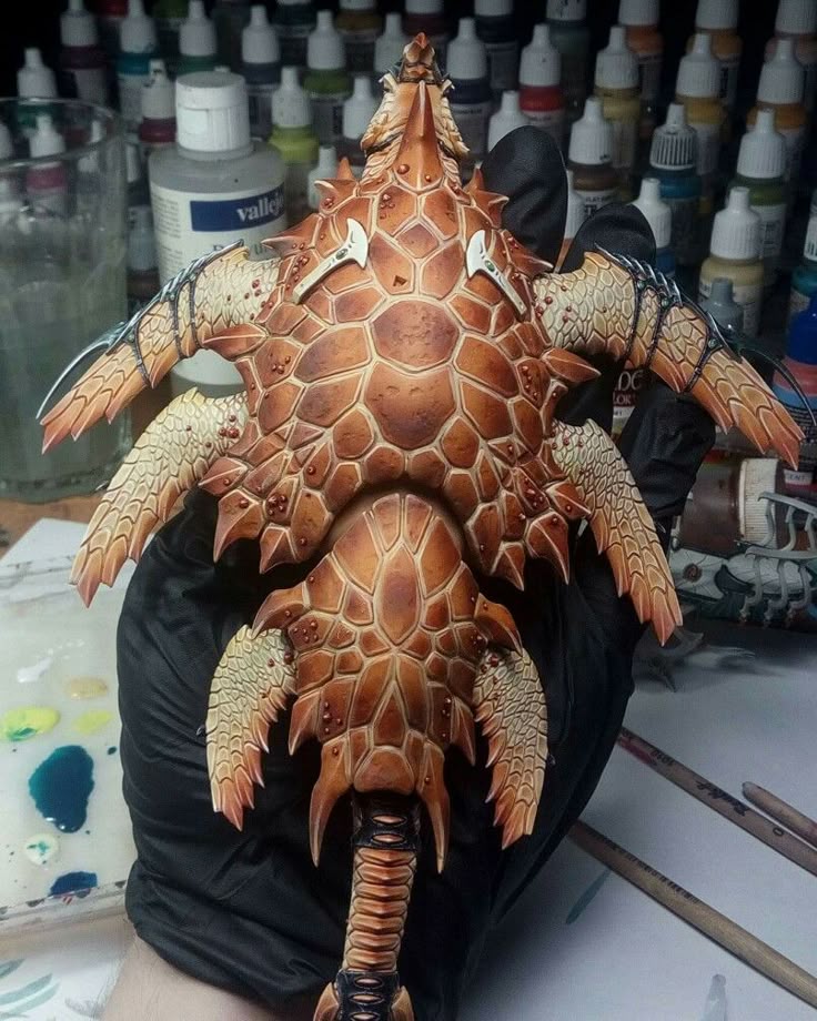 the image shows a detailed, handpainted miniature figure held in a gloved hand. the figure appears to be a fantasy creature with a carapace of large, brown and orange scales, adorned with sharp spines and smaller feathery appendages. the figure is set against a background filled with various paint bottles and miniature painting supplies, suggesting it's a hobby project related to tabletop games or wargaming. the level of detail and shading indicates a skilled painter.