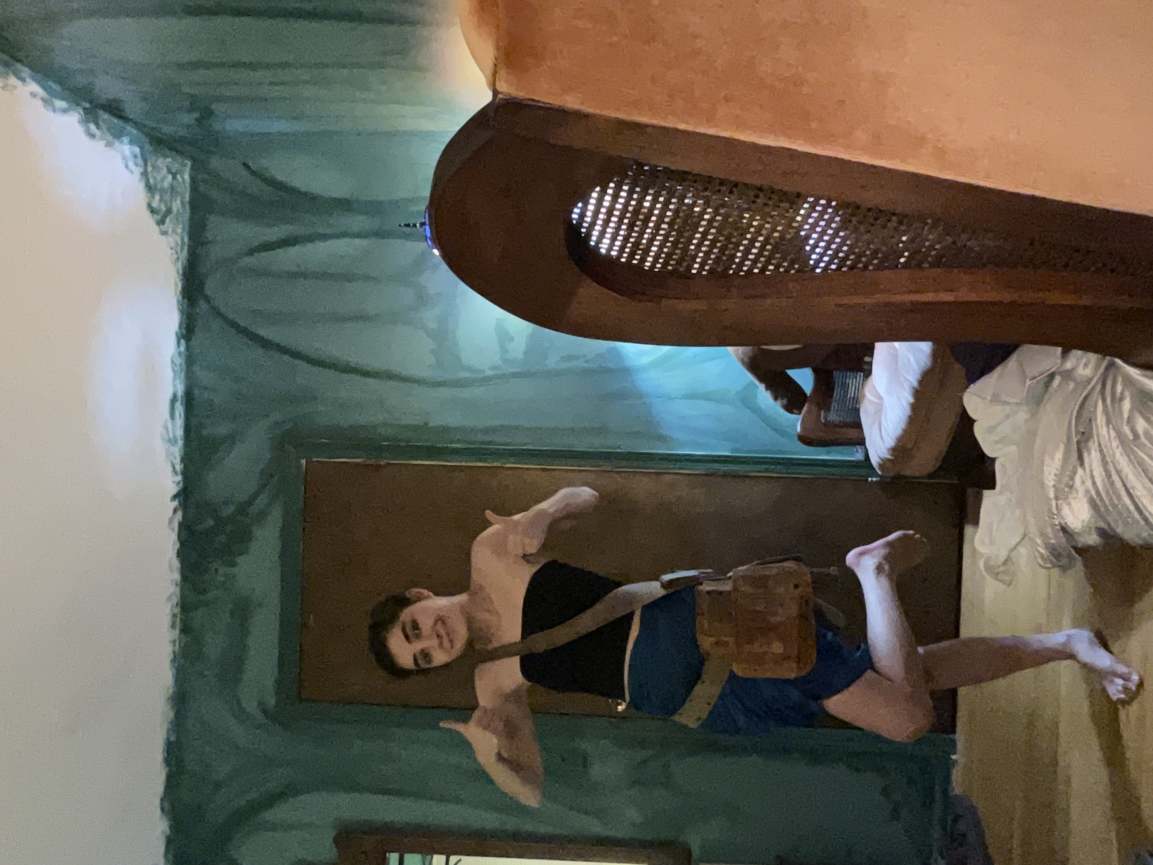 a young woman with short dark hair is standing in a room with teal painted walls, giving a thumbs up with both hands. she's wearing a short blue skirt, a brown shoulder and waist bag, and is barefoot.  she is smiling. the room has some unique furniture, including an orange chair and a wooden chair with a woven back.  the walls have a textured, uneven paint job.