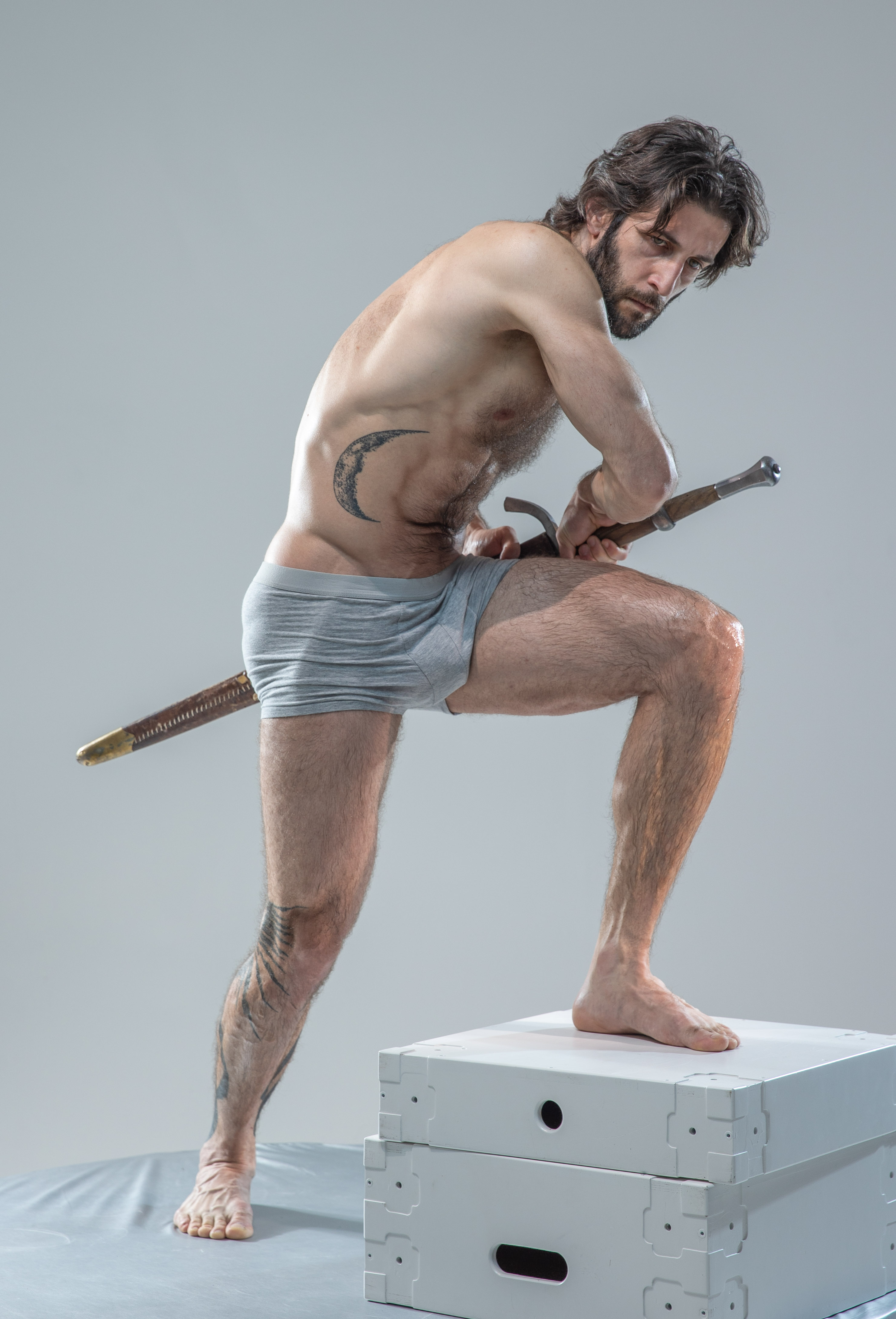 a shirtless, muscular man with long dark hair and a beard stands in a dynamic pose on top of two white boxes. he is wearing grey underwear and holds a sword in his hands, the tip pointing towards the left. he has a tattoo on his left hip. the background is a light grey. the overall image gives a sense of strength and power.
