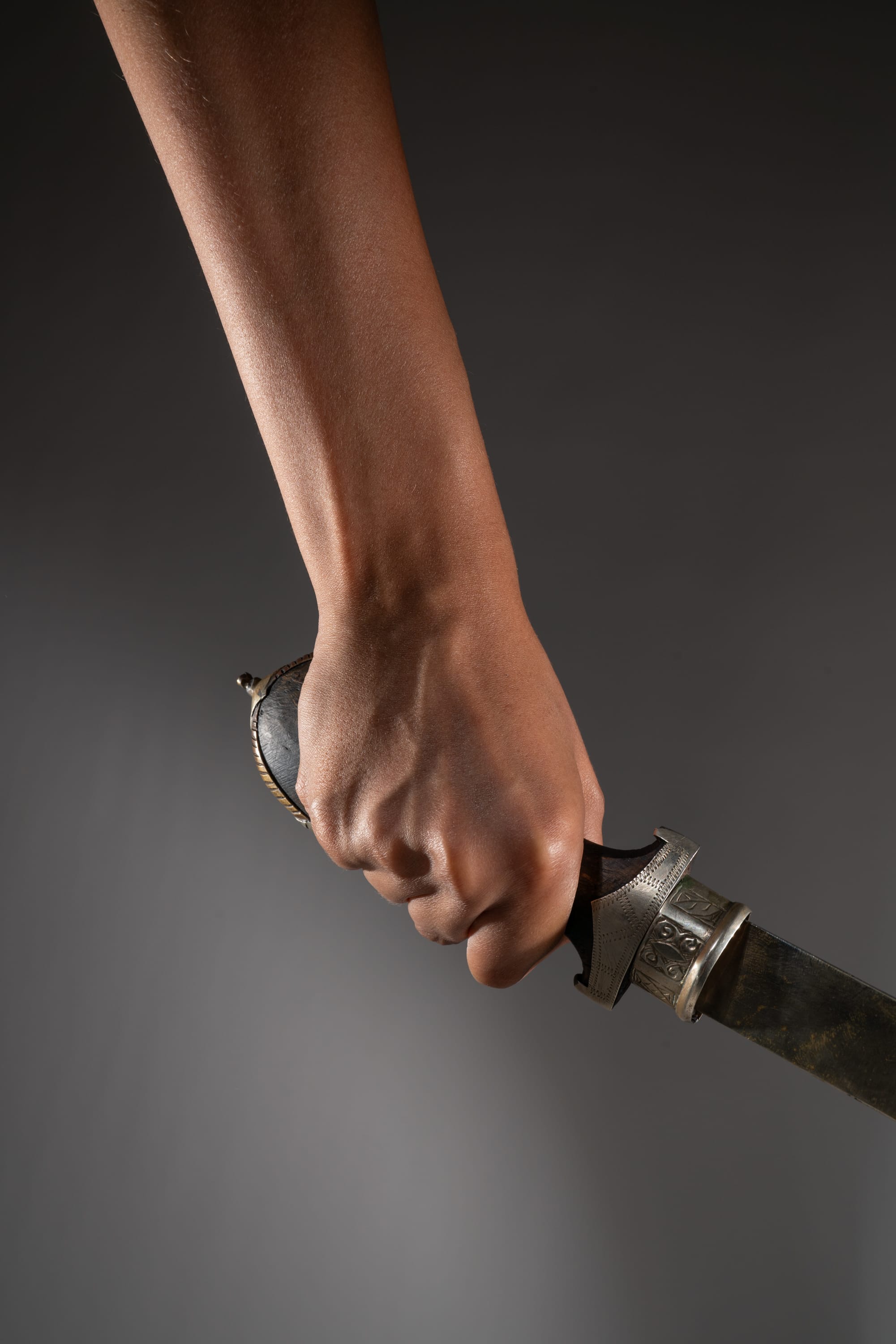 a closeup shot of a hand gripping the ornate handle of a sword against a dark gray background. the hand is tensed, highlighting the potential for both power and danger associated with the weapon. the sword's blade extends out of the frame, adding to the sense of mystery and threat.