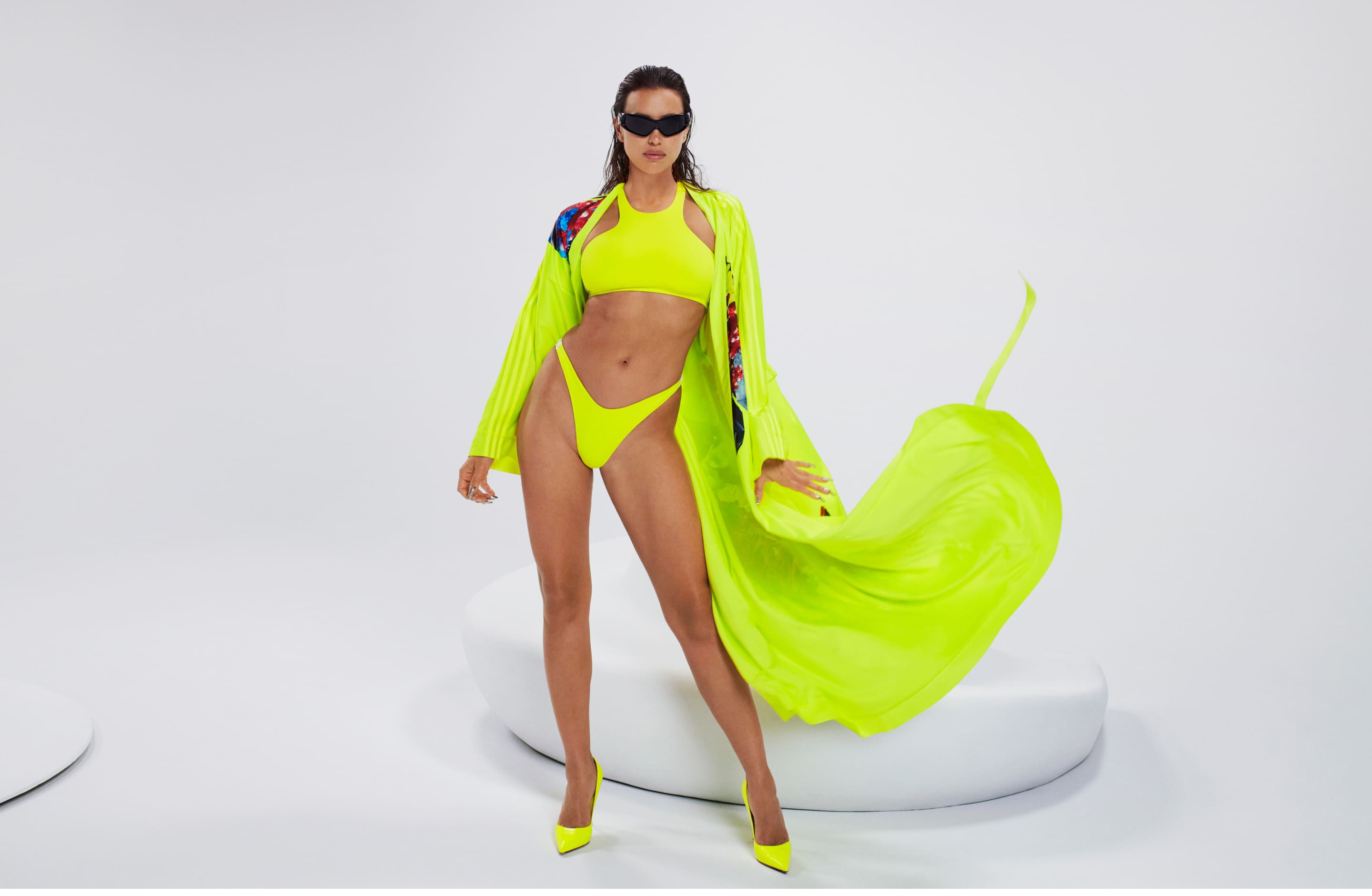 a model in a yellow neon swimsuit and  matching robe  stands with one leg raised, holding the robe in the air, while the other leg is bent and her foot is flat on the ground.  she is wearing sunglasses and neon yellow heels. her long dark hair is straight and falls to her shoulders. she is looking directly at the camera. the background is a white studio wall and the floor is white.  the image is shot from a low angle.