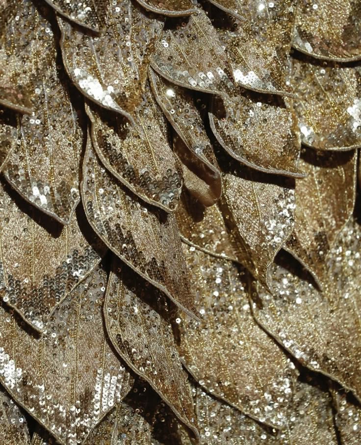 a closeup image shows a pattern of golden, leafshaped fabric pieces covered in sequins. the leaves overlap each other, creating a textured and shimmering surface. the overall effect is luxurious and decorative.