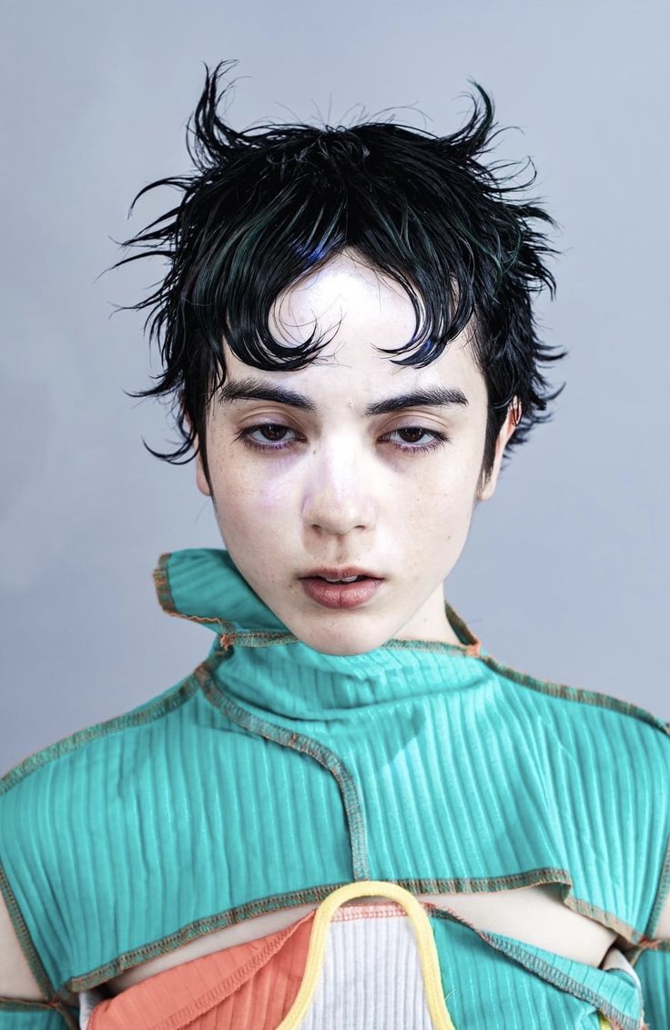 a closeup portrait of a woman with short, wet hair, dressed in a blue cut out shirt, looking directly at the camera. her face is clean with minimal makeup and her expression is serious. she is posing in a studio against a neutral grey background.