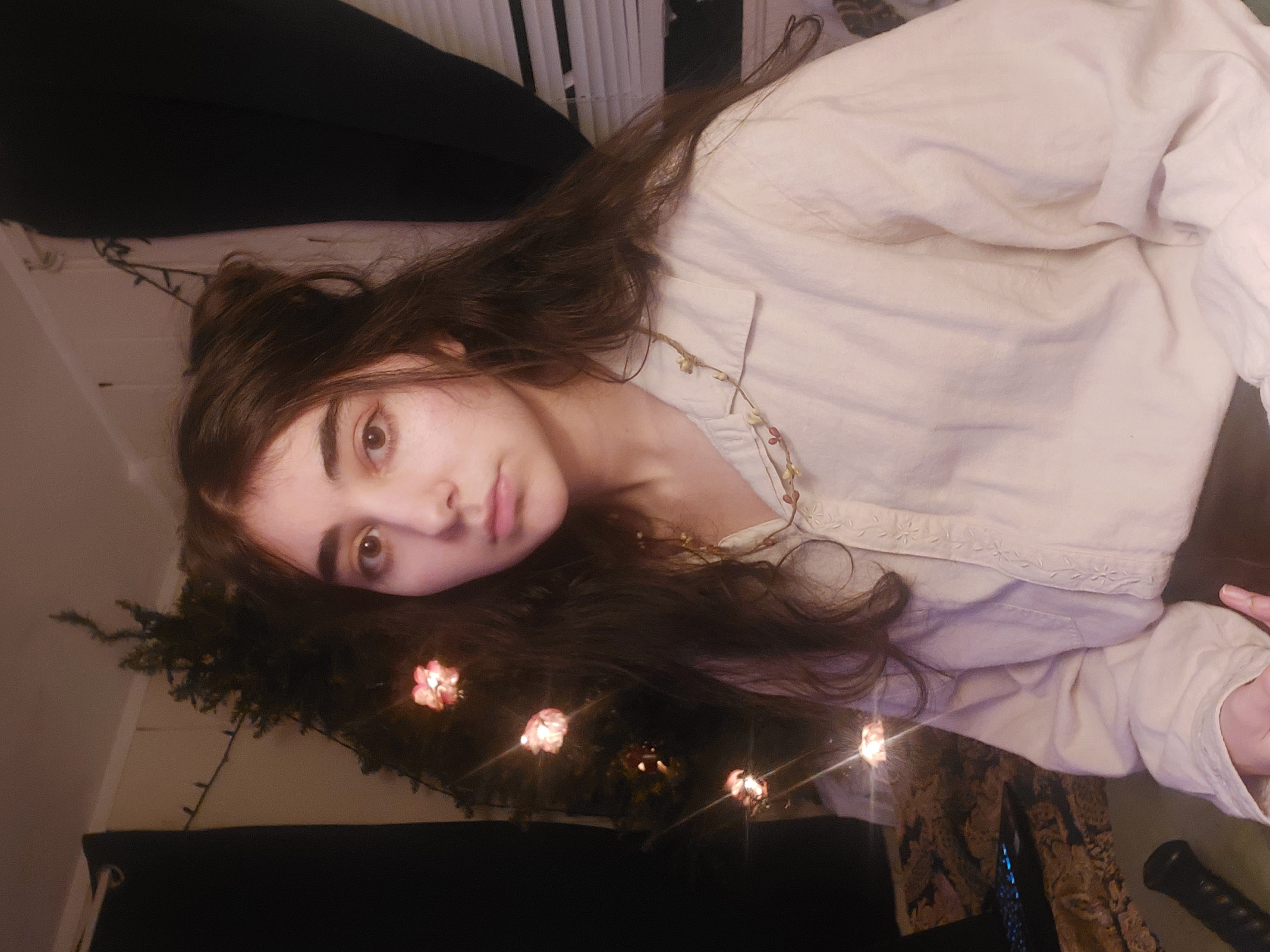 a closeup portrait of a young woman with long brown hair partially styled in a halfupdo. she's wearing a light beige, longsleeved buttondown shirt and a delicate flower necklace.  the setting appears to be indoors, with a christmas tree decorated with lights visible in the background. the lighting is soft and dim, creating a warm and intimate atmosphere. the woman's expression is serious and neutral.