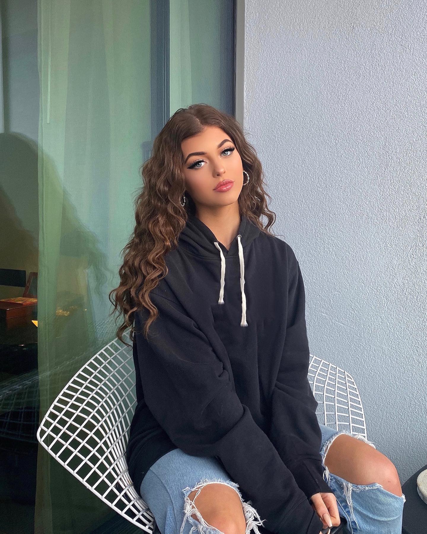 a young woman with long, curly brown hair is sitting in a white, gridpatterned chair outdoors. she's wearing a black hoodie with white drawstrings and ripped light blue jeans. her makeup is visible, including winged eyeliner and pink lipstick. she has blue eyes and is wearing hoop earrings. the background is a plain gray wall and a green, reflective glass surface. the lighting appears to be natural.