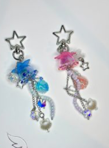 two adorable handmade keychains, crafted with resin bellflowers, shimmering beads, and delicate faux pearls, dangle gracefully from silver starshaped clasps. the left keychain showcases a captivating blue bellflower, while the right keychain features a charming pink one. each keychain is adorned with a mesmerizing assortment of iridescent beads, adding a touch of sparkle and whimsy.