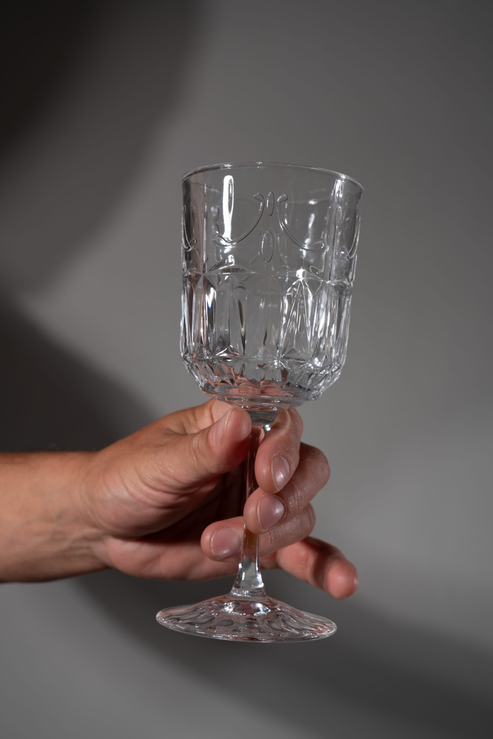 a man's hand holds a clear, empty glass with a decorative pattern. the background is a solid gray, with shadows cast from offcamera lighting.