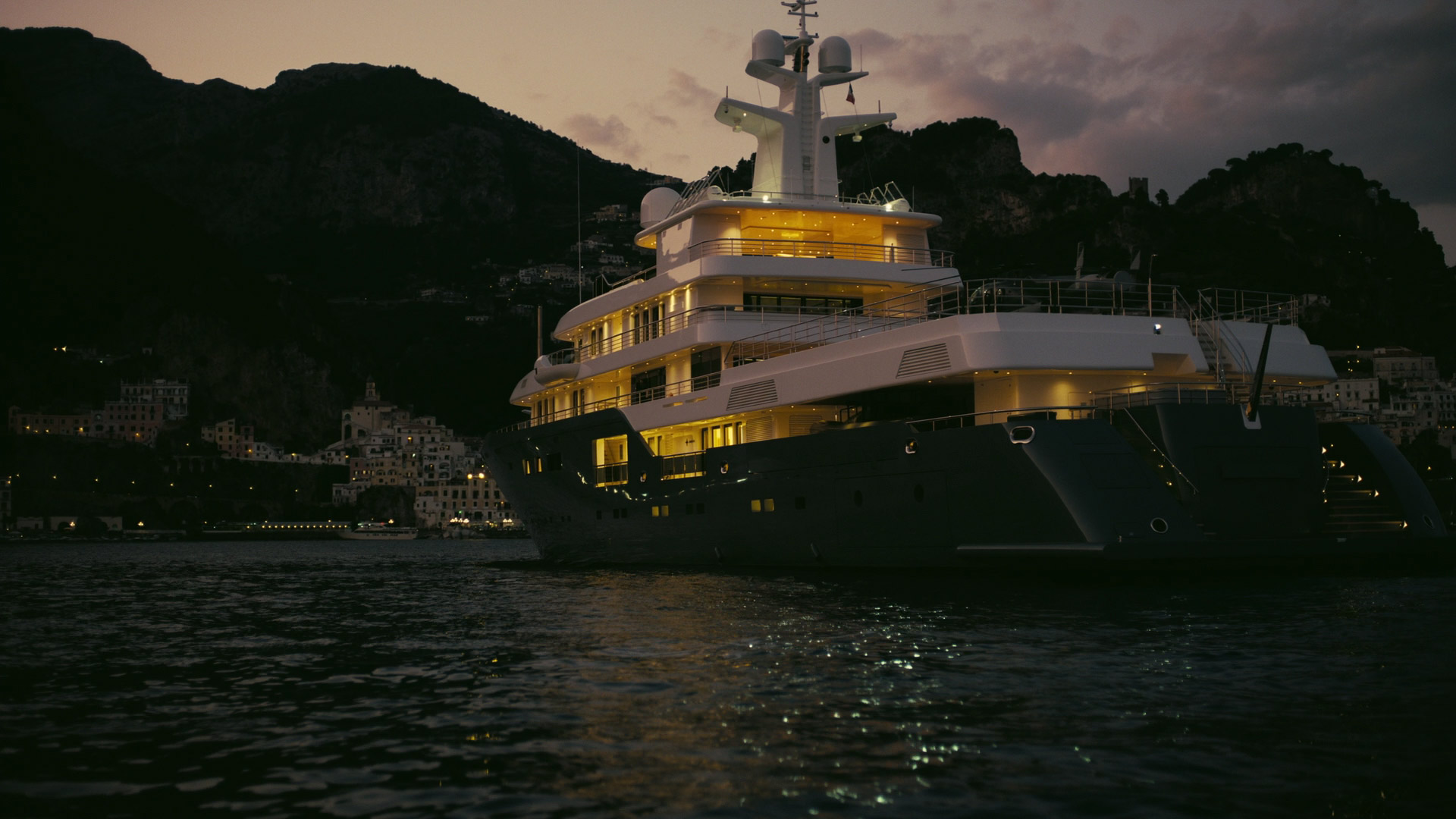 a large, luxurious yacht is shown at night, lit up by warm, artificial lighting. it is facing away from the viewer and to the left, and is in front of a dark, hilly shoreline dotted with lights from houses. the water is dark and reflects the lights from the yacht and shoreline. the sky is a dark, purplishblue.