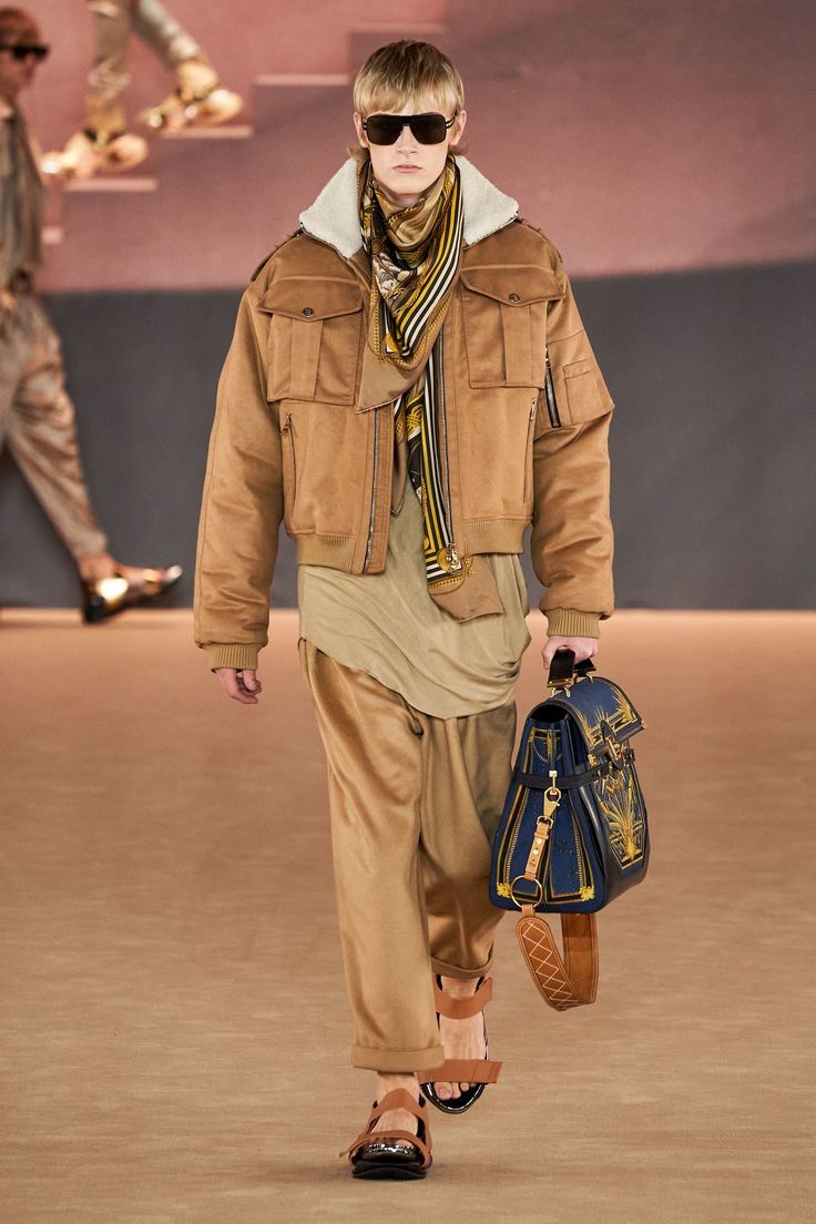 a male fashion model walks down a runway showcasing a fall/winter collection. he's wearing a tan suede jacket with a sherpa collar, a beige shirt, matching pants, brown sandals, and a patterned scarf.  he carries a dark blue and gold shoulder bag. the model is wearing sunglasses and has short blond hair.  the background is blurred, showing a tancolored runway and another person walking in the distance. the overall style is contemporary and elegant.