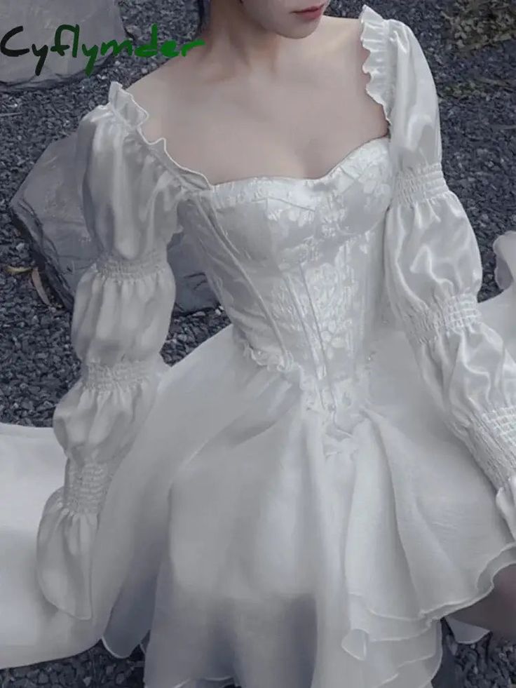 a woman is shown wearing a short white dress with a corset bodice and puff sleeves. the dress has a square neckline, a cinched waist, and a ruffled skirt. the bodice appears to have a subtle floral pattern. the sleeves are made of a slightly shiny material, possibly satin, and are gathered at the wrists with elastic. the skirt appears to be made of a sheer fabric, possibly chiffon.