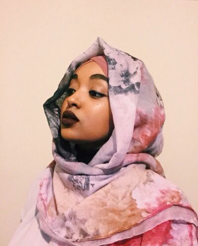 a portrait of a woman wearing a hijab. the hijab is made of a beautiful floral pattern with shades of pink, purple and grey. she is looking at the camera with a soft gaze. her makeup is done subtly with a soft lip color and defined eyes. the background is a plain white wall.
