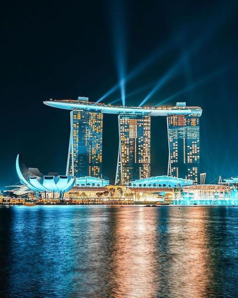 the image showcases the iconic marina bay sands hotel in singapore at night.  three towering hotel towers are connected by a distinctive curved rooftop structure.  the buildings are brilliantly illuminated, with multiple spotlights creating dramatic light beams that reach up into the dark sky. the calm waters of marina bay reflect the city lights, creating a mesmerizing mirror image of the cityscape.  the artscience museum is visible in the foreground, adding to the scene's vibrancy. the overall effect is a breathtaking display of modern architecture and stunning nighttime illumination.