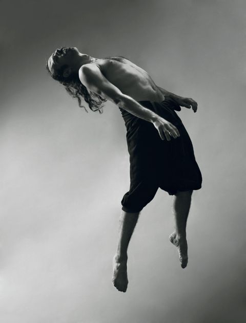 a black and white image of a man in midair, he is suspended in the air and looks as if he is dancing.  the man is barefooted and his body is in a graceful and athletic pose.  the background is a plain wall.