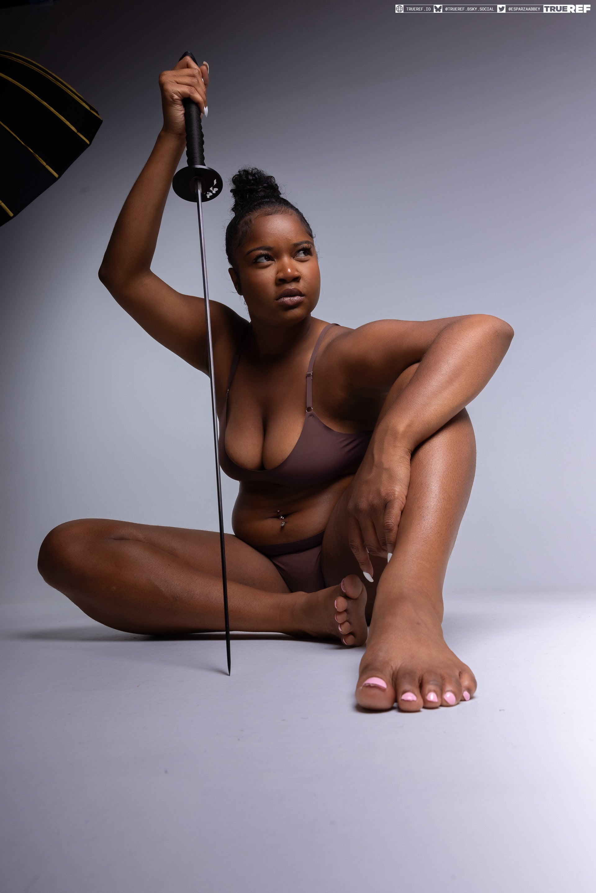 a studio shot of a darkskinned woman in brown underwear sitting crosslegged on a gray surface, holding a sword with the blade pointing downwards. she has her hair in a bun and is looking to the right, with her feet visible in the foreground.