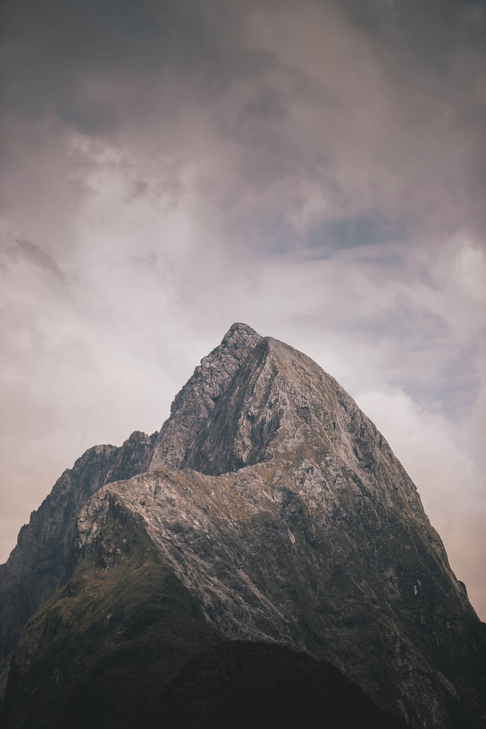 a majestic mountain peak dominates the frame, its rugged, rocky slopes rising sharply into the overcast sky. the muted, earthy tones of the mountain contrast subtly with the soft grays and blues of the clouds, creating a sense of both power and tranquility.  the low angle shot emphasizes the peak's imposing height and grandeur.