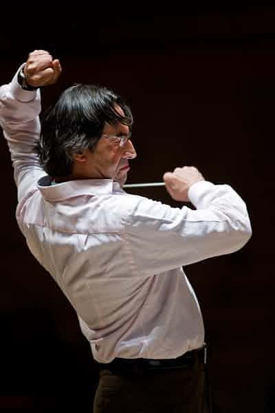 a conductor in a white shirt and black pants is conducting an orchestra. he is holding a baton in his right hand and is focused on the music. the conductor is on stage, performing in front of a dark background. the image is taken during a concert or performance. the lighting is focused on the conductor.