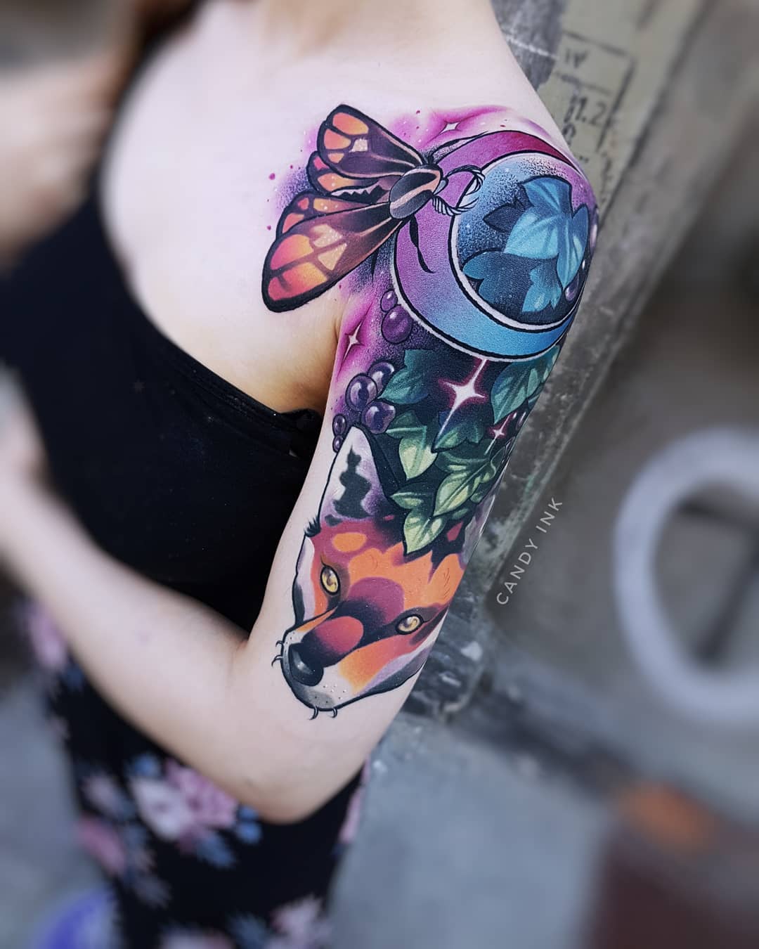 a closeup photograph showcasing a colorful tattoo on a woman's upper arm and shoulder. the tattoo features a realistic fox head, a stylized moth in flight, a crescent moon adorned with grapes and leaves, and scattered stars, all rendered in a vibrant neotraditional style.