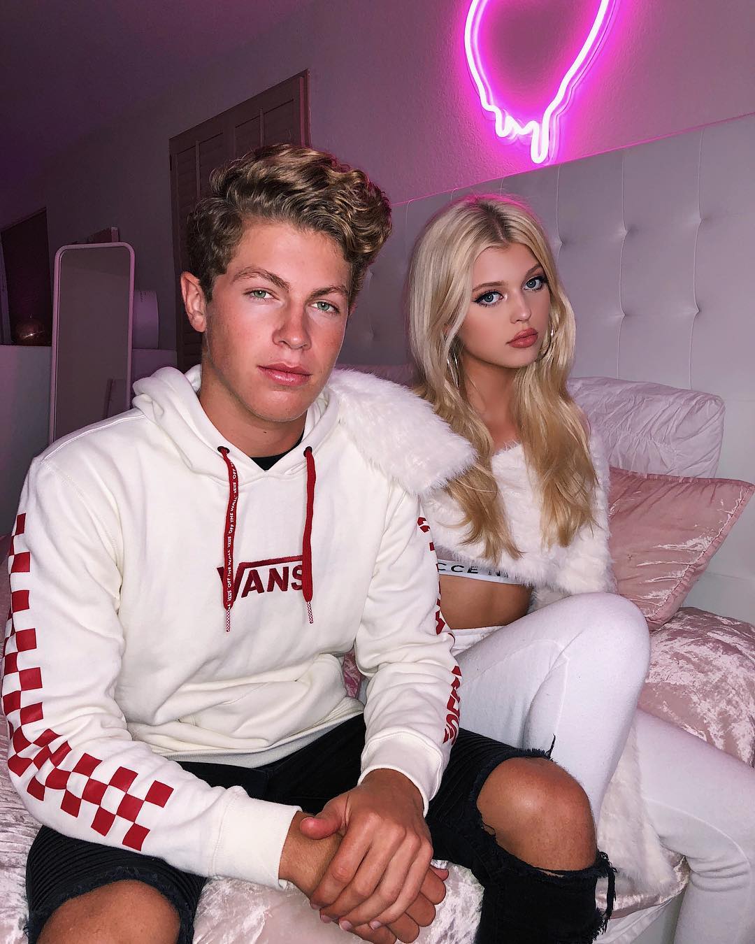 a photo of a young man and a young woman sitting together on a bed. the man is wearing a white hoodie with the word "vans" on the front and red checkered sleeves, along with black ripped jeans. the woman is wearing a white top and white pants. behind them is a headboard and a pink neon light.