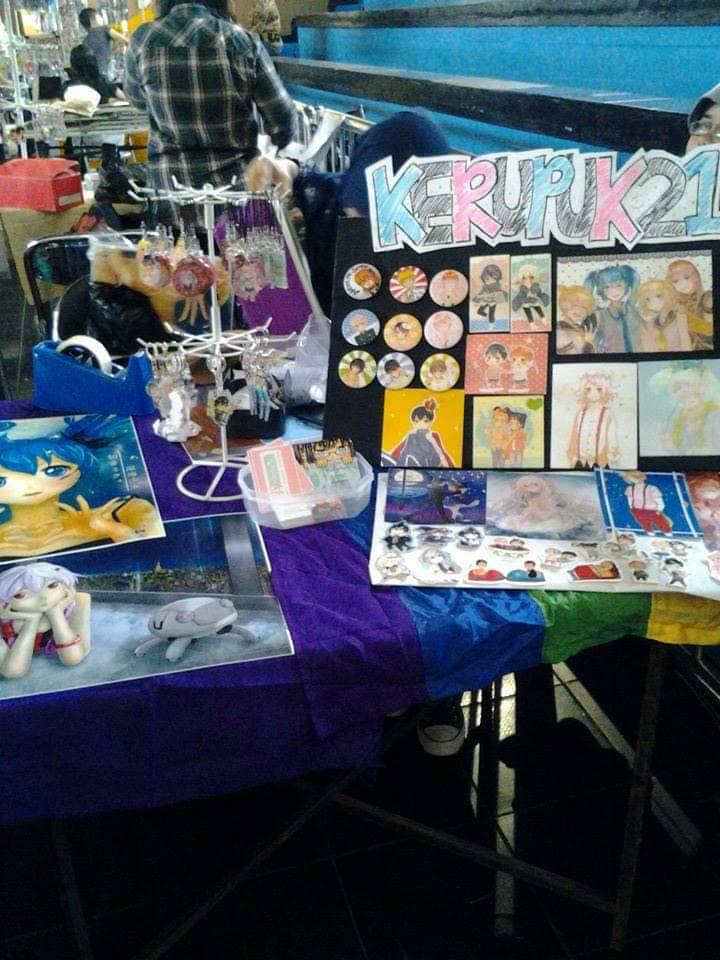 this image shows a table set up for selling merchandise at an anime convention. the table is covered in a purple and blue tablecloth with rainbow stripes. there are anime and manga character pins and badges on display, as well as some other items. the table is set up in front of a white wall and there are people in the background. there is a sign on the table that says "kerupuk," which is a type of indonesian snack. the table is decorated with various anime and mangathemed items, suggesting that the convention is focused on japanese pop culture.