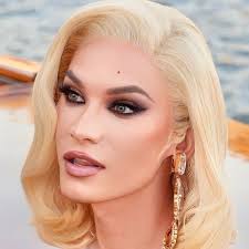 the image is a closeup portrait of a blonde woman with wavy, shoulderlength hair.  she's wearing glamorous makeup, including noticeable eyeshadow, and long, dangling golden earrings.  the background is blurry but suggests an outdoor setting possibly near water, with a hint of a boat visible. the overall impression is one of elegance and beauty.  the woman's expression is calm and slightly pensive.