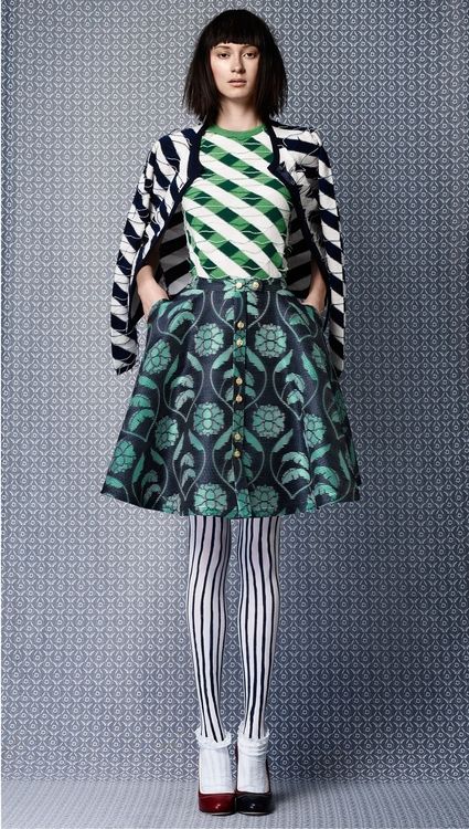 a full shot of a fashionable young woman. she is wearing a teal and green floral buttondown skirt, a green and white striped top, a black and white striped cardigan, white and black striped tights, and black heels. she has a bob haircut and is posing in front of a patterned background.