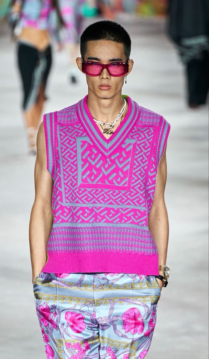 a male fashion model walks the runway wearing a vibrant pink sweater vest with a geometric pattern and light blue floral patterned pants. he accessorizes with pink sunglasses, a necklace, and bracelets. the overall look is summery and fashionable with a blend of bright and pastel colors. the background is blurred, focusing attention on the model.