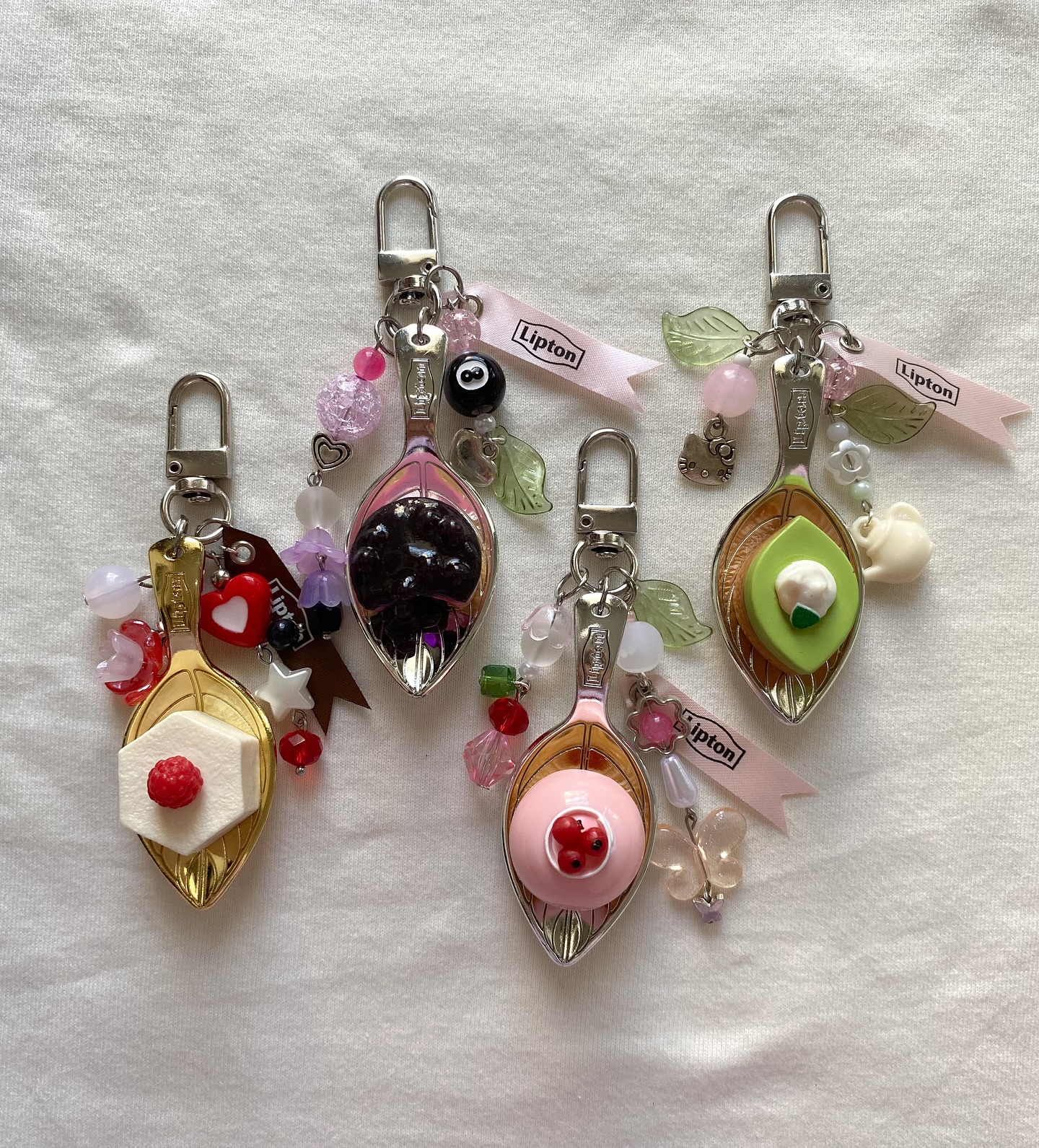 the image shows a collection of four decorative keychains lying on a white background. each keychain features a spoon filled with a different "food" item, such as a miniature avocado, blackberry, raspberry, or a sugar cube with a raspberry on top. they are all attached to a keychain clip and are decorated with an assortment of beads, charms, and ribbons that add to their whimsical and handmade appearance. there are different charms like a teapot, hello kitty, butterfly, and beads of different shapes like hearts, leaves and flowers. the ribbons have "lipton" printed on them.