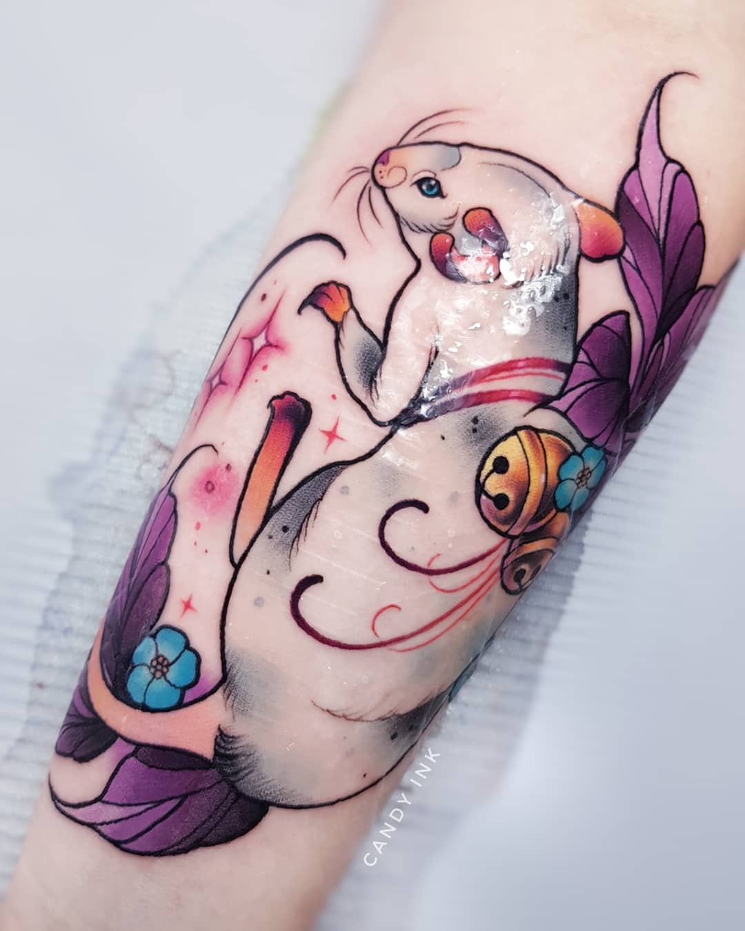 a neotraditional style tattoo of a rat on someone's arm. the rat is white with pink details, and there are purple flowers and orange and yellow swirls around it.