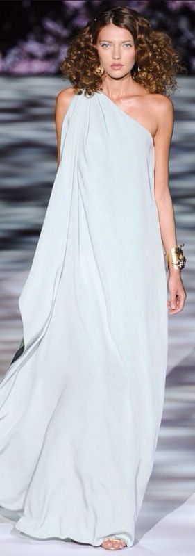 a female model walks the runway in a stunning white one shoulder dress with long, flowing fabric. her hair is styled in loose curls, and she accessorizes with gold jewelry, including a large bracelet on her wrist. her makeup is soft and natural, complementing her elegant look.  the overall image conveys a sense of sophistication, beauty, and glamor.
