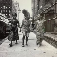a black and white photo of three young girls happily holding hands and walking down a city sidewalk. they are all wearing dresses and smiling. the photo evokes a sense of joy, friendship, and childhood innocence.
