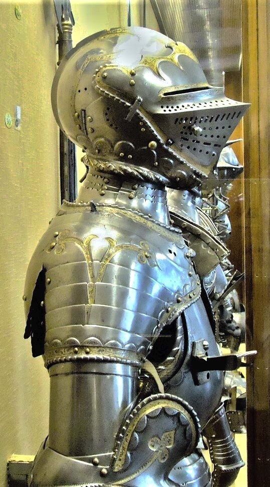 a closeup view of an ornate suit of medieval armor displayed in a museum. the armor is made of polished steel, featuring intricate details and embellishments. the helmet, with its raised visor, is particularly striking. the lighting is artificial, typical of a museum setting, highlighting the metallic sheen and craftsmanship of the armor.