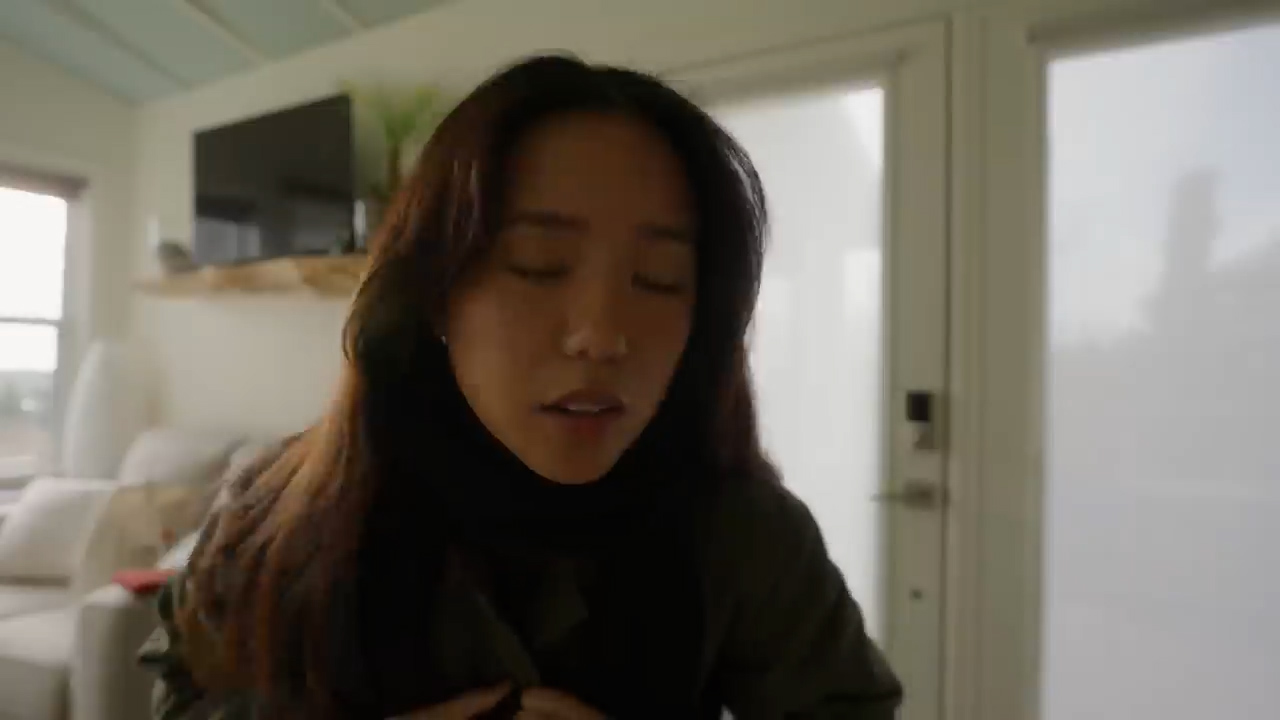 a young asian woman with long dark hair and wearing a green jacket is shown from the chest up. she is inside a house, and the lighting suggests it's daytime with natural light coming through a window. the woman is looking down with her eyes closed, and her expression appears contemplative or thoughtful.