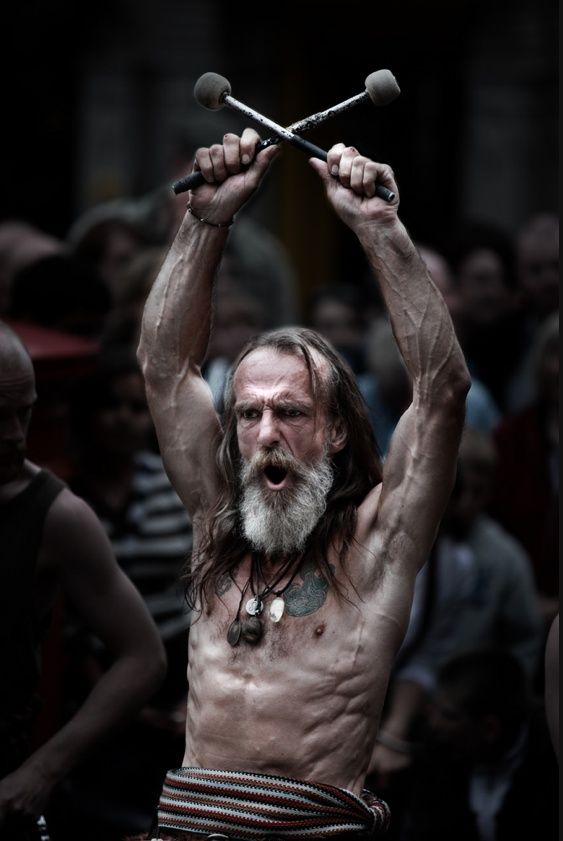 a shirtless bearded man with long white hair is holding two drumsticks above his head and shouting. he is surrounded by a crowd of people watching his performance. he is a street performer  and he is giving an energetic performance.
