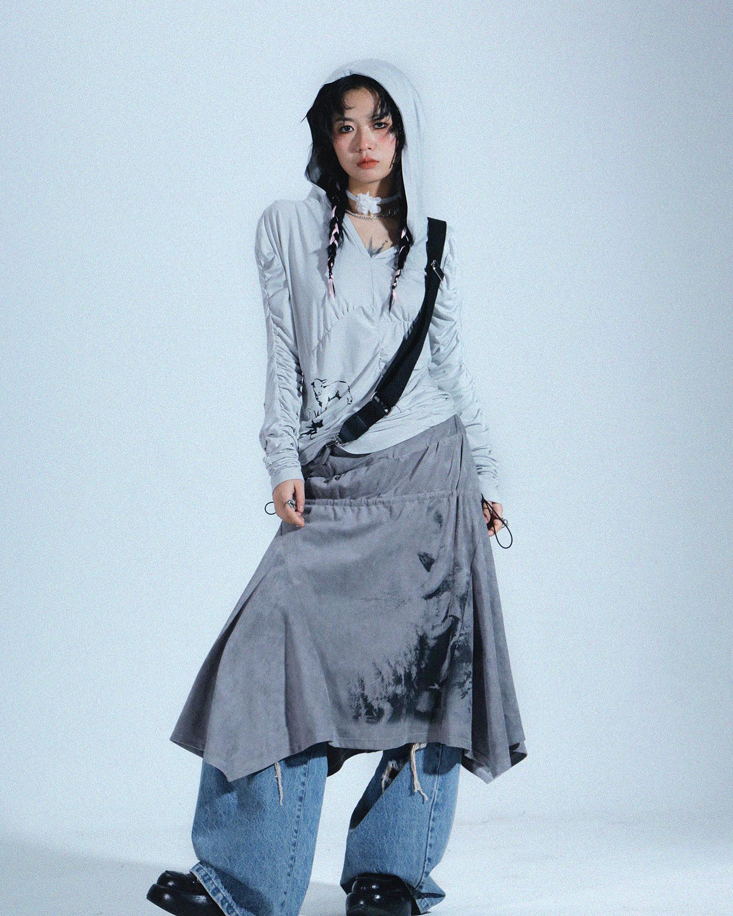 a full shot of a fashion model posing in a studio against a light grey background. she is wearing a light grey hooded longsleeve top with an artistic print, paired with an asymmetrical, light grey skirt featuring a dark inklike splatter print.  the skirt is layered over distressed light blue jeans.  she is accessorized with a choker necklace and a black crossbody strap. her overall style is streetwearinspired, blending grunge elements with contemporary fashion. the pose and lighting create a stylish and cool image that showcases the outfit's design.