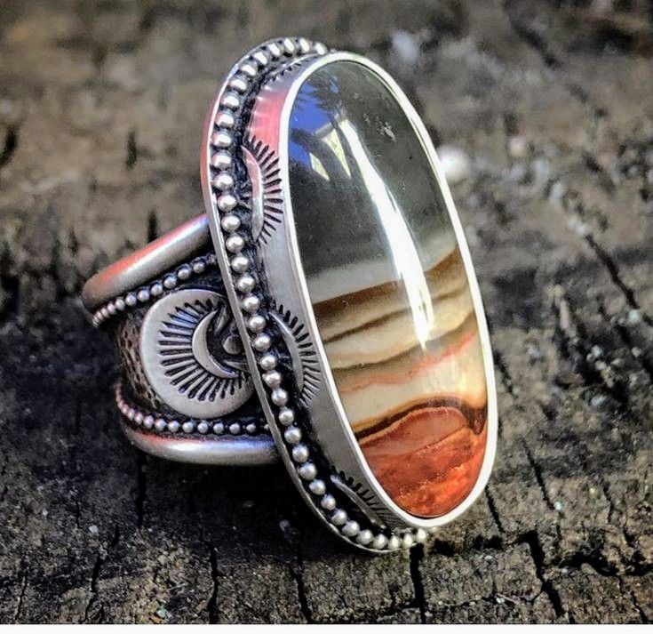 a closeup shot of a beautiful handmade silver ring with an oval agate stone. the band of the ring is adorned with a southwest design and the stone is a mix of browns, tans, and reds with a beautiful dark grey band going down the middle. the ring is sitting on a rough wooden surface.